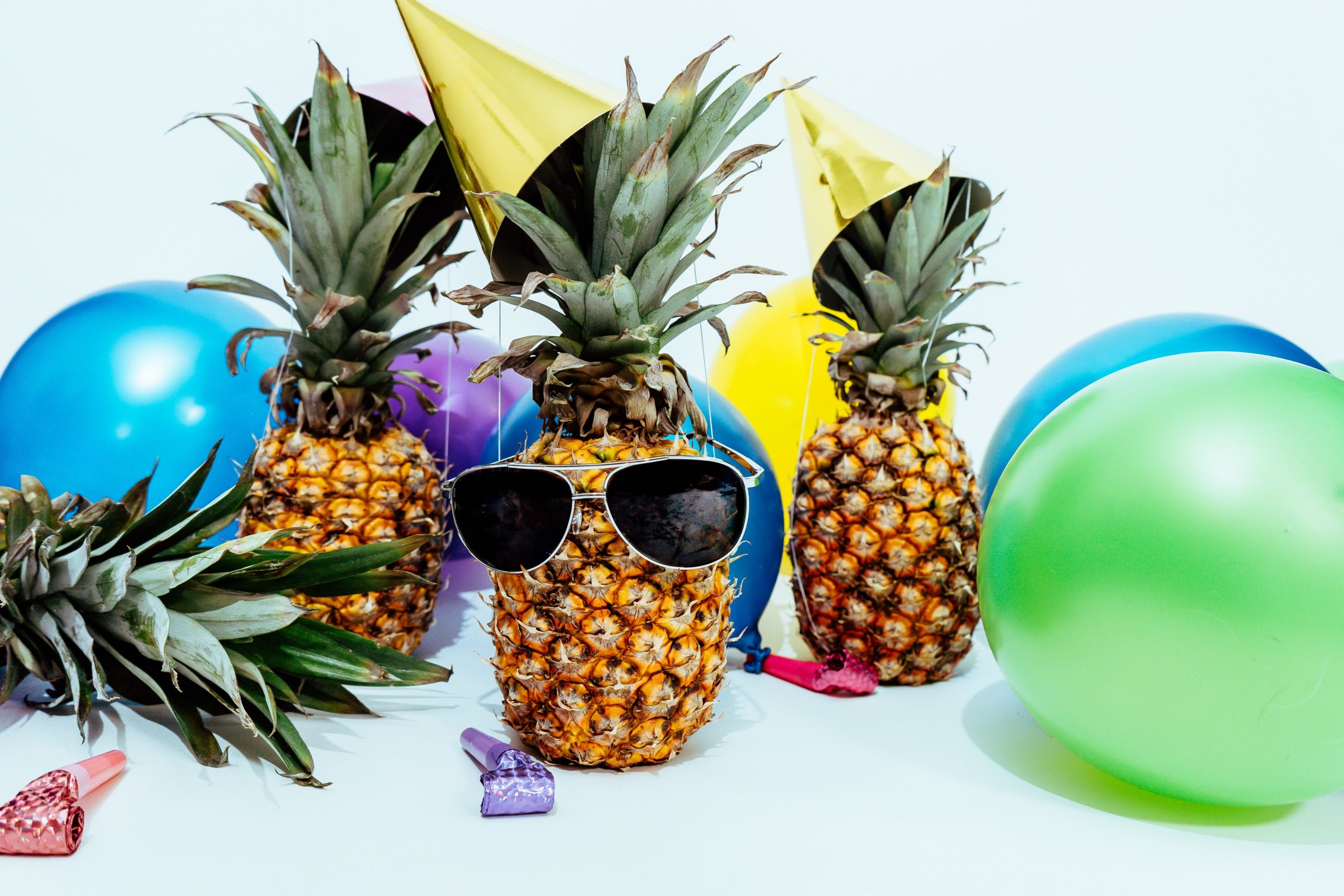 Ananas sa sunčanim naočalama i žutom party kapicom, okružen šarenim balonima i party zviždaljkama.